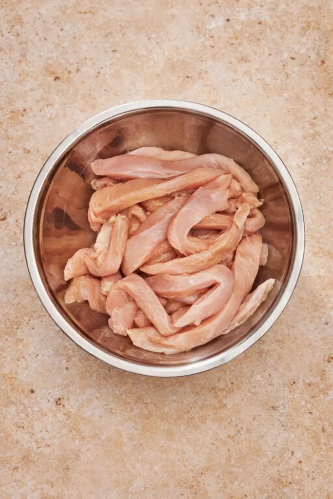 Fresh raw chicken breast strips sliced into thin "fries" sitting in a stainless steel mixing bowl on a tan stone countertop.