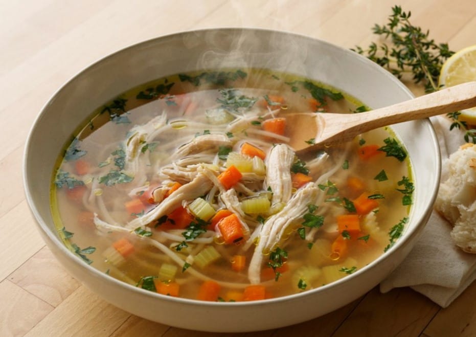 Grandma's Chicken Soup Recipe: The Simple Bowl 7 Traditional Grandma's Chicken Soup served in a plate with a spoon inside, accompanied by lemon wedges and fresh green herbs beside the dish.