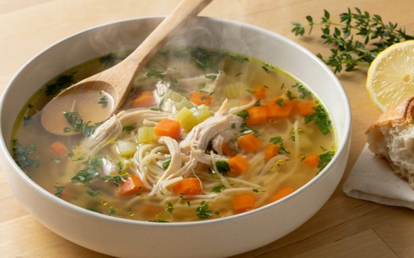 Grandma's Chicken Soup Recipe: The Simple Bowl 6 Homemade Grandma's Chicken Soup in a shallow plate with a spoon resting inside, garnished with lemon slices and fresh greens on the sid