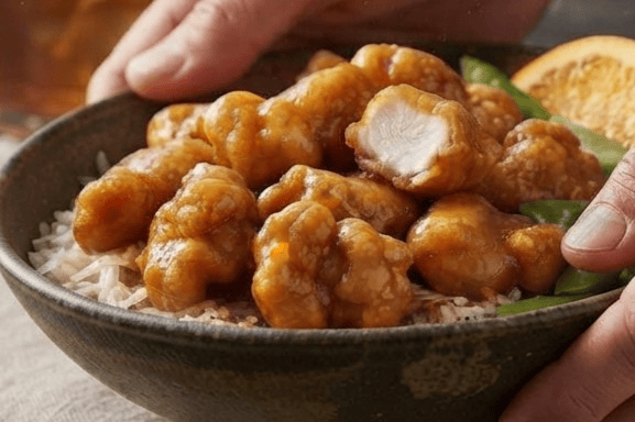 A rustic bowl of orange chicken, rice, and snap peas is held by two hands. The chicken pieces are glazed and golden, with one piece cut in half to reveal the meat. Green snap peas and an orange slice provide contrast. The setting is a dark table with a linen placemat, and a glass of iced tea is in the background